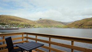 A wooden deck with a table and chairs overlooking a lake with a town and hills in the background. - Luxurious Waterfront Retreat | Spectacular Views (Vestmanna)