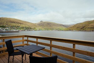A wooden deck with a table and chairs overlooking a lake with a town and hills in the background.