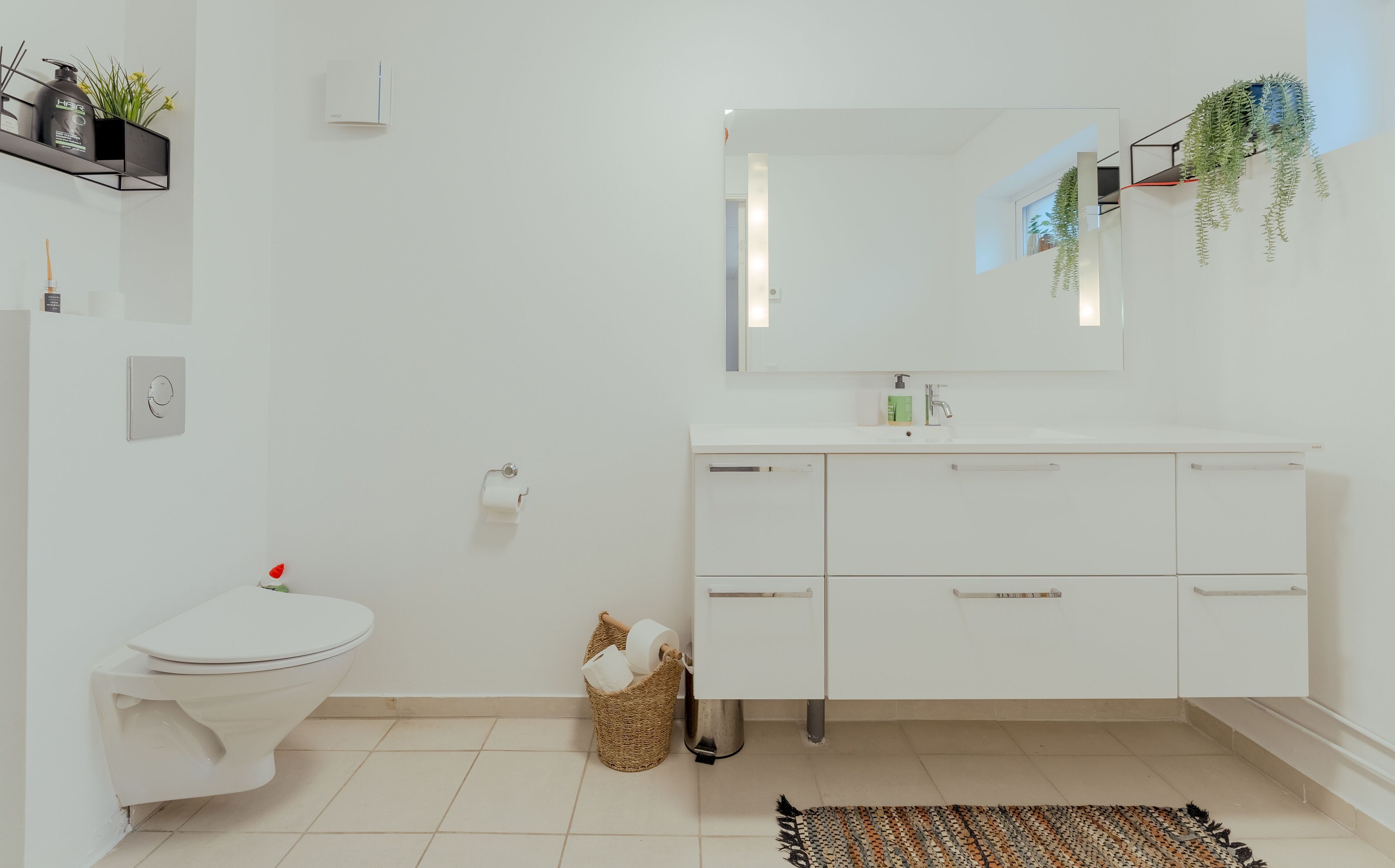 A modern bathroom with a toilet, sink, and mirror. There is a basket of toiletries and a plant on the wall.