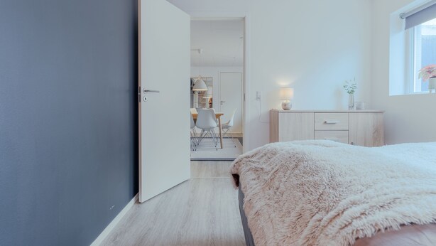 A modern bedroom with a fluffy white bed, a wooden dresser, and a desk with chairs.