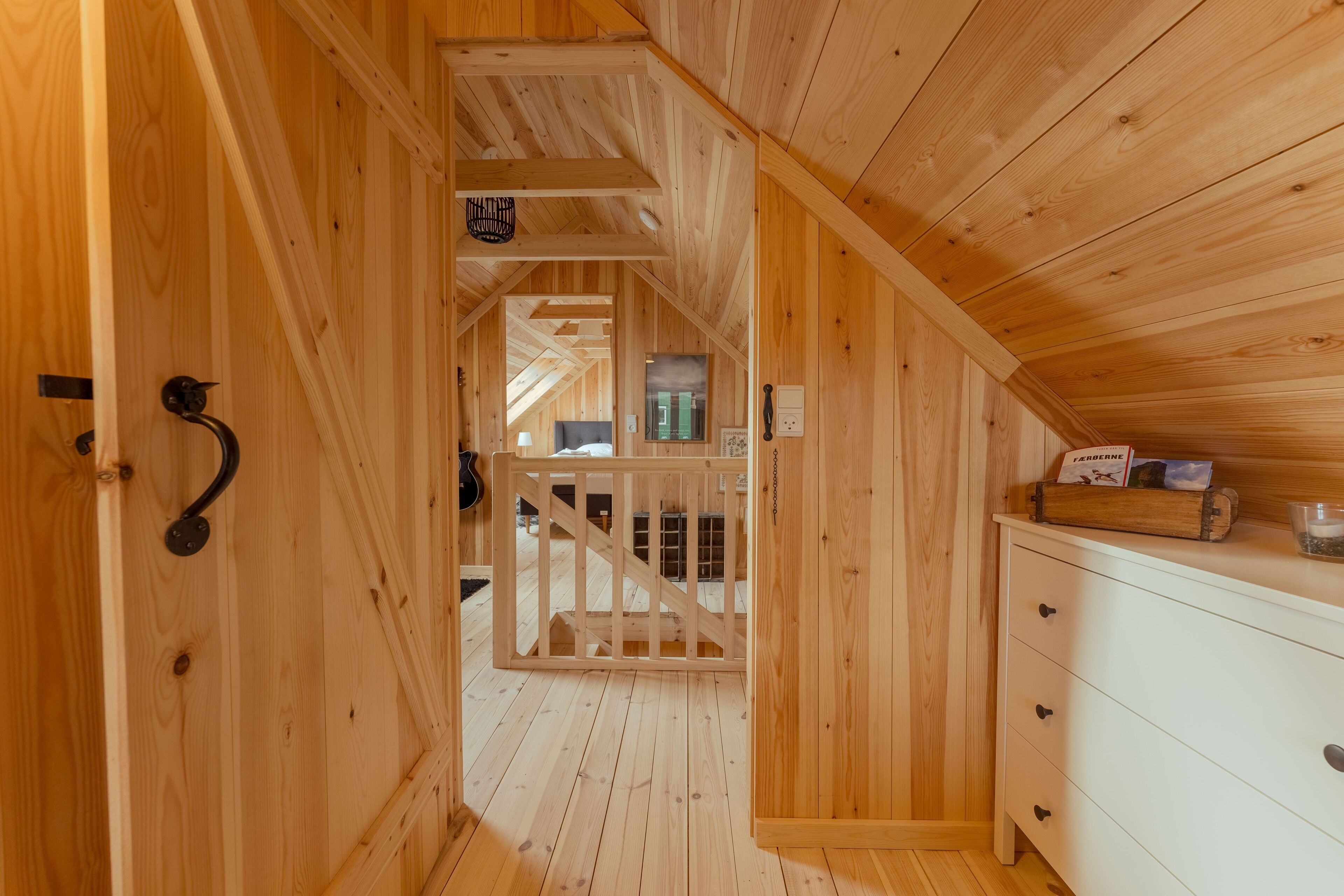 Un intérieur en bois avec un escalier menant à un niveau supérieur, une commode blanche et une petite étagère sur laquelle sont posés divers objets.