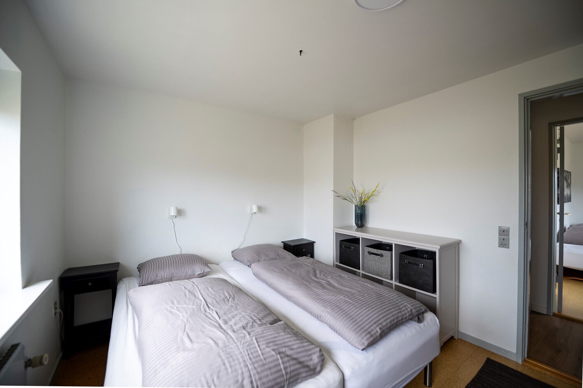 Quarto de hotel moderno com duas camas, um guarda-roupa branco, uma pequena mesa de cabeceira e um criado-mudo.