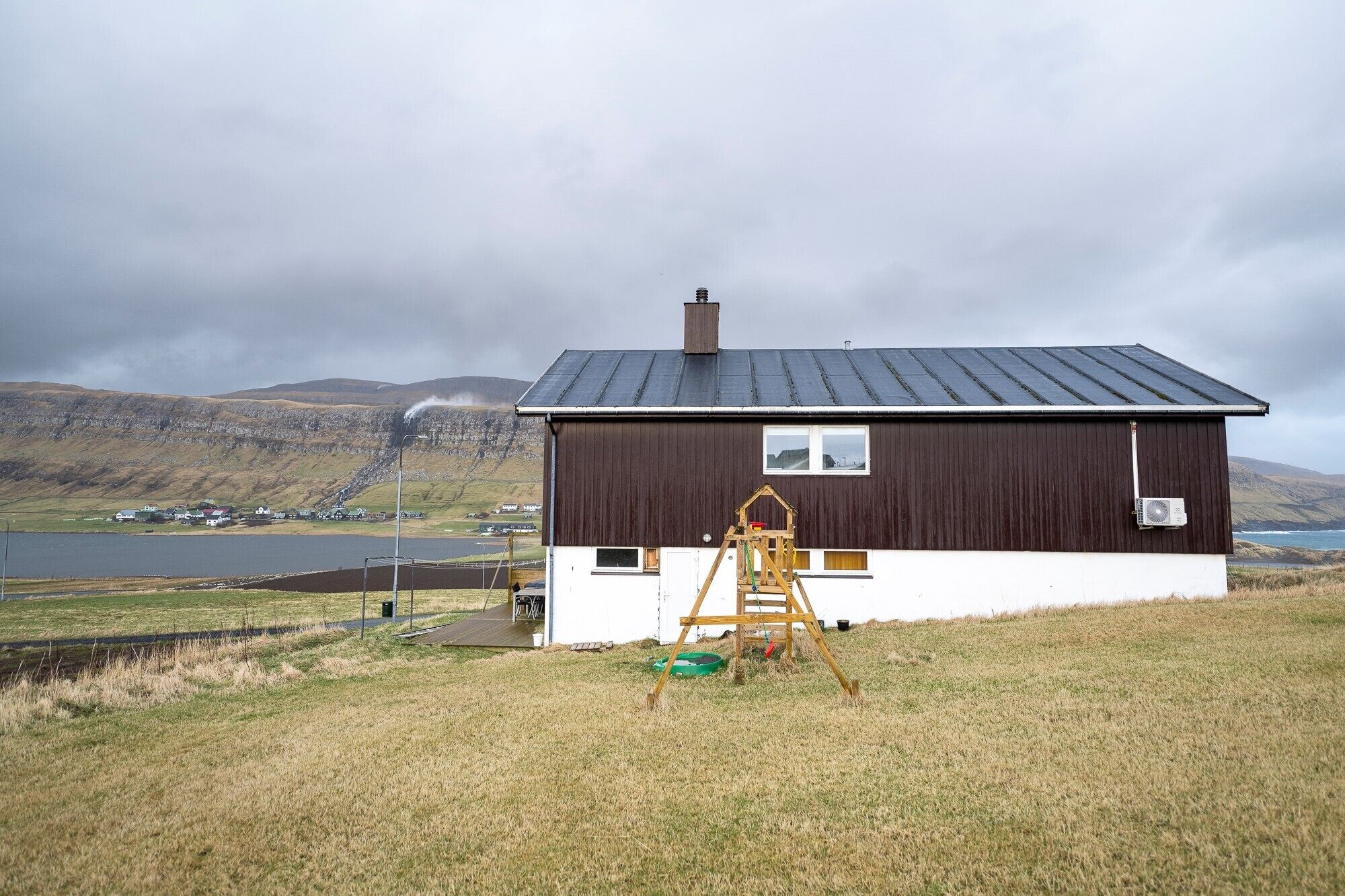 A single-story house with a dark brown exterior and white trim, situated on a grassy hill with a view of a lake and mountains in the background.