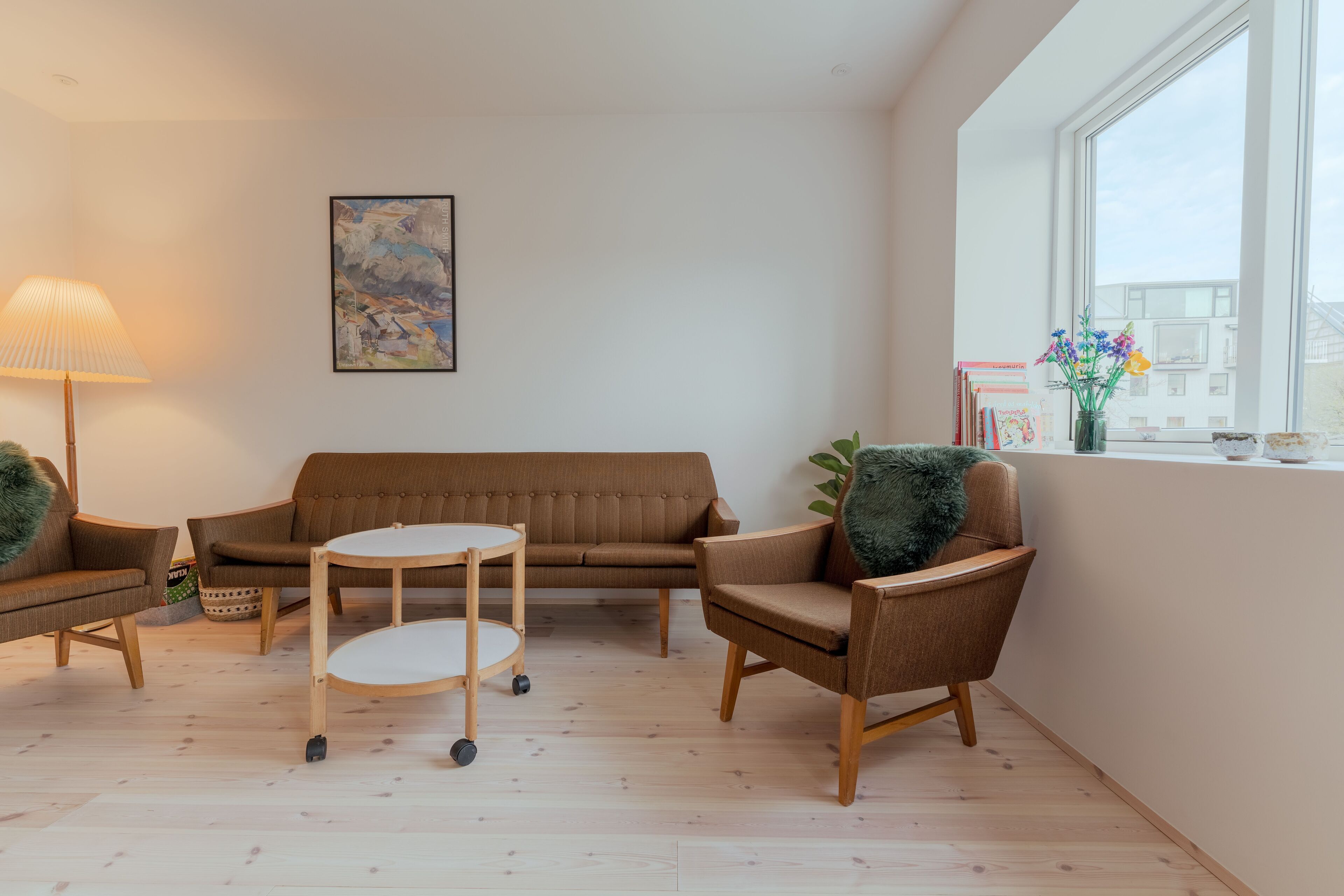 Een moderne woonkamer met een bruine bank, twee fauteuils, een klein rond tafeltje en een lamp. Er is een raam met uitzicht op gebouwen en een lijst met een afbeelding aan de muur.