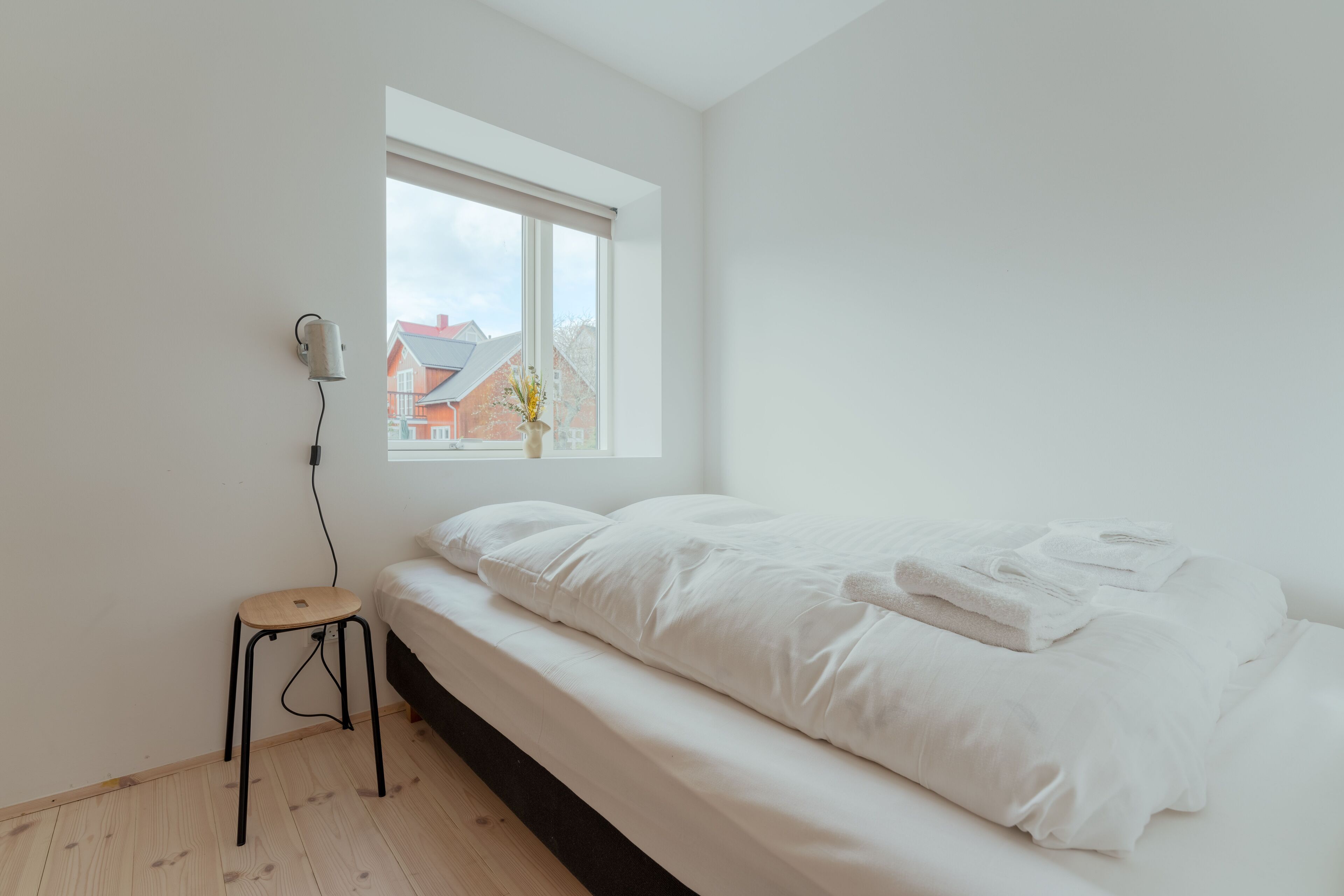 Camera da letto minimalista con un letto singolo, uno sgabello e una finestra che si affaccia su edifici.