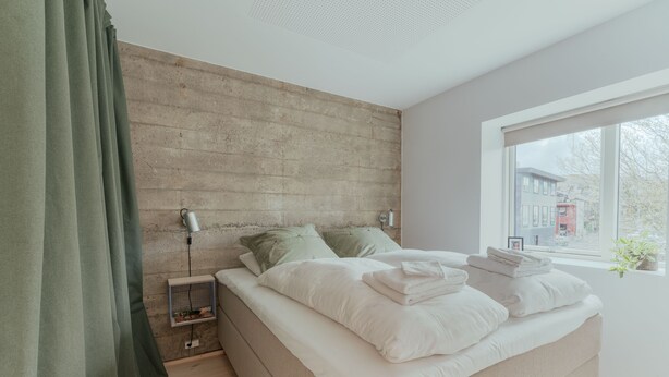 A bedroom with a bed, green curtains, a wall-mounted heater, and a window with a view of buildings. - Charming Central Tórshavn Retreat - 2Br Apartment (Tórshavn)
