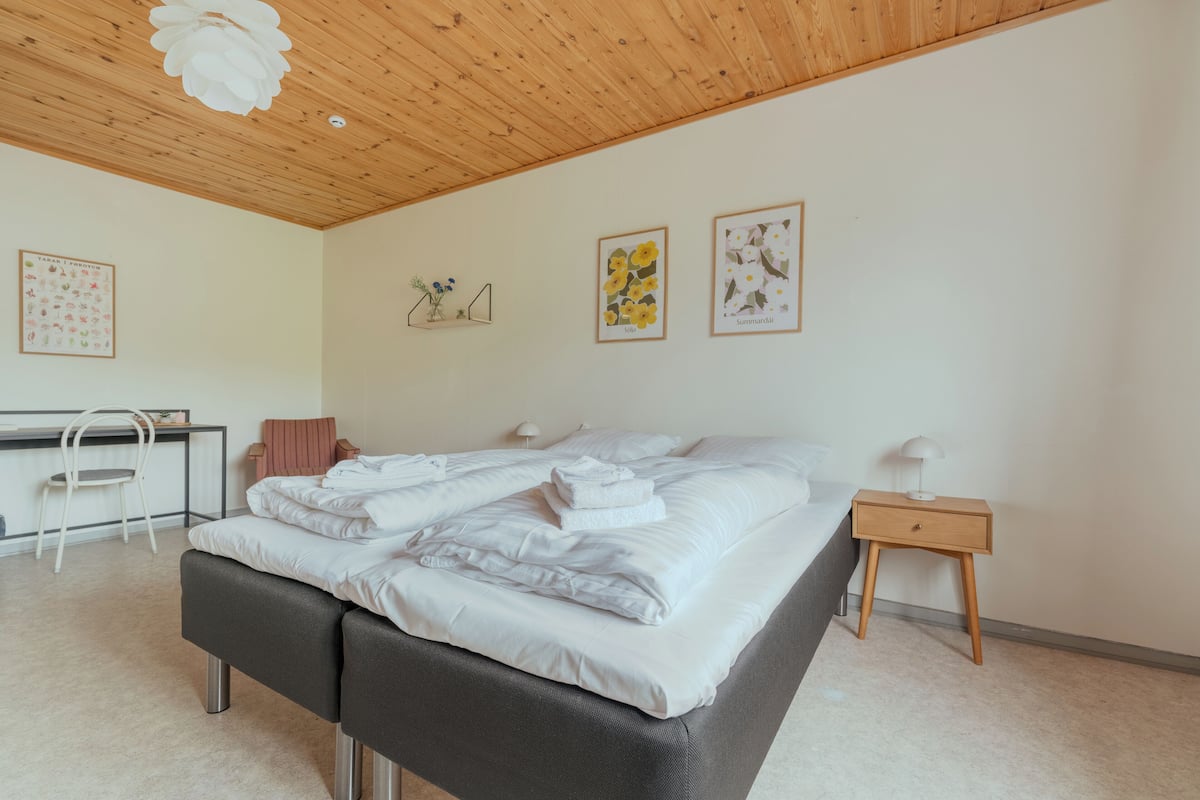 Een netjes opgemaakt bed met witte lakens, een houten nachtkastje met een lamp, een bureau met een stoel, en twee ingelijste botanische prints aan de muur.