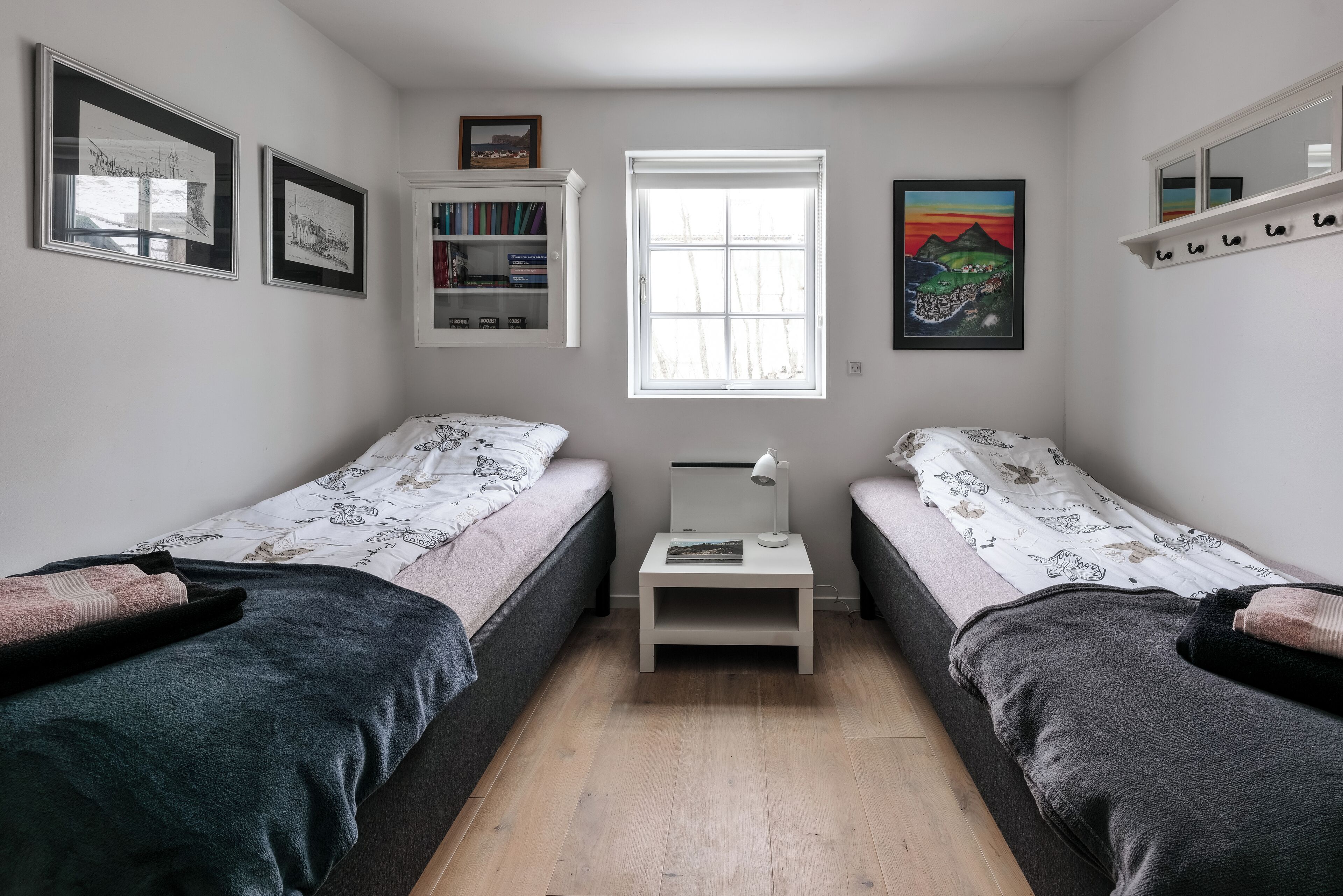 Une chambre avec deux lits simples, une table de chevet avec une lampe, une bibliothèque et une fenêtre.