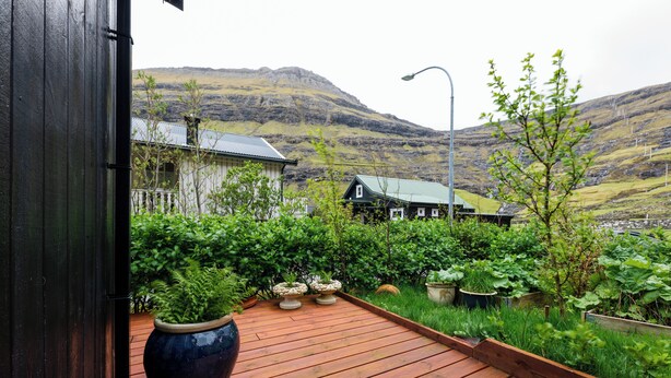 Una terraza de madera con plantas en macetas, vistas a casas y un paisaje montañoso.