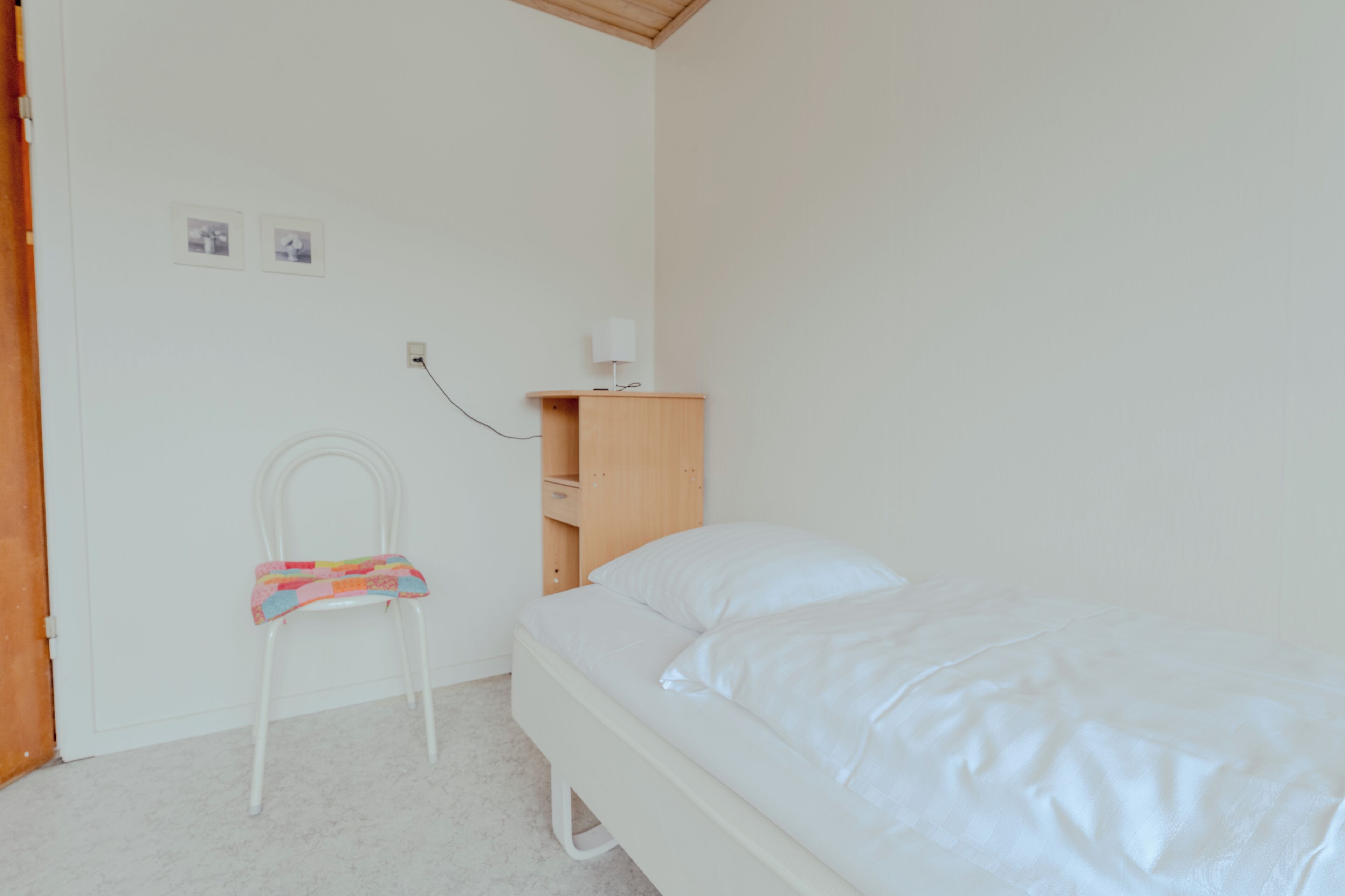 A small, clean room with a single bed, a chair, a nightstand, and a wall socket.
