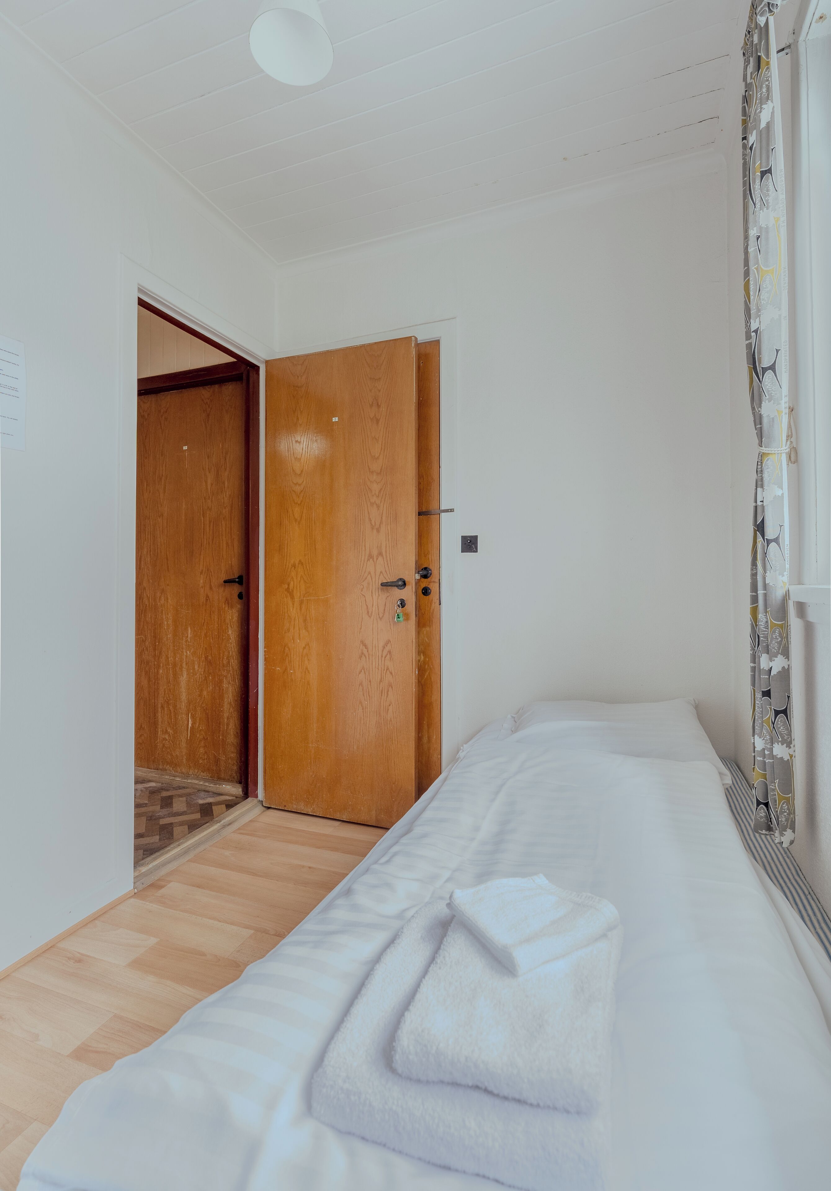 A bedroom with a wooden door, a bed with white bedding, and a window with a curtain.