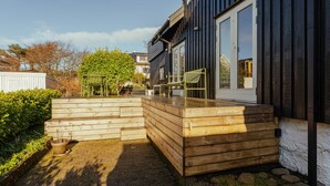 A wooden deck with chairs, a potted plant, and a stone pathway leading to a house with a dark exterior and large windows. - Charming Retreat In The Heart Of Tórshavn (Tórshavn)