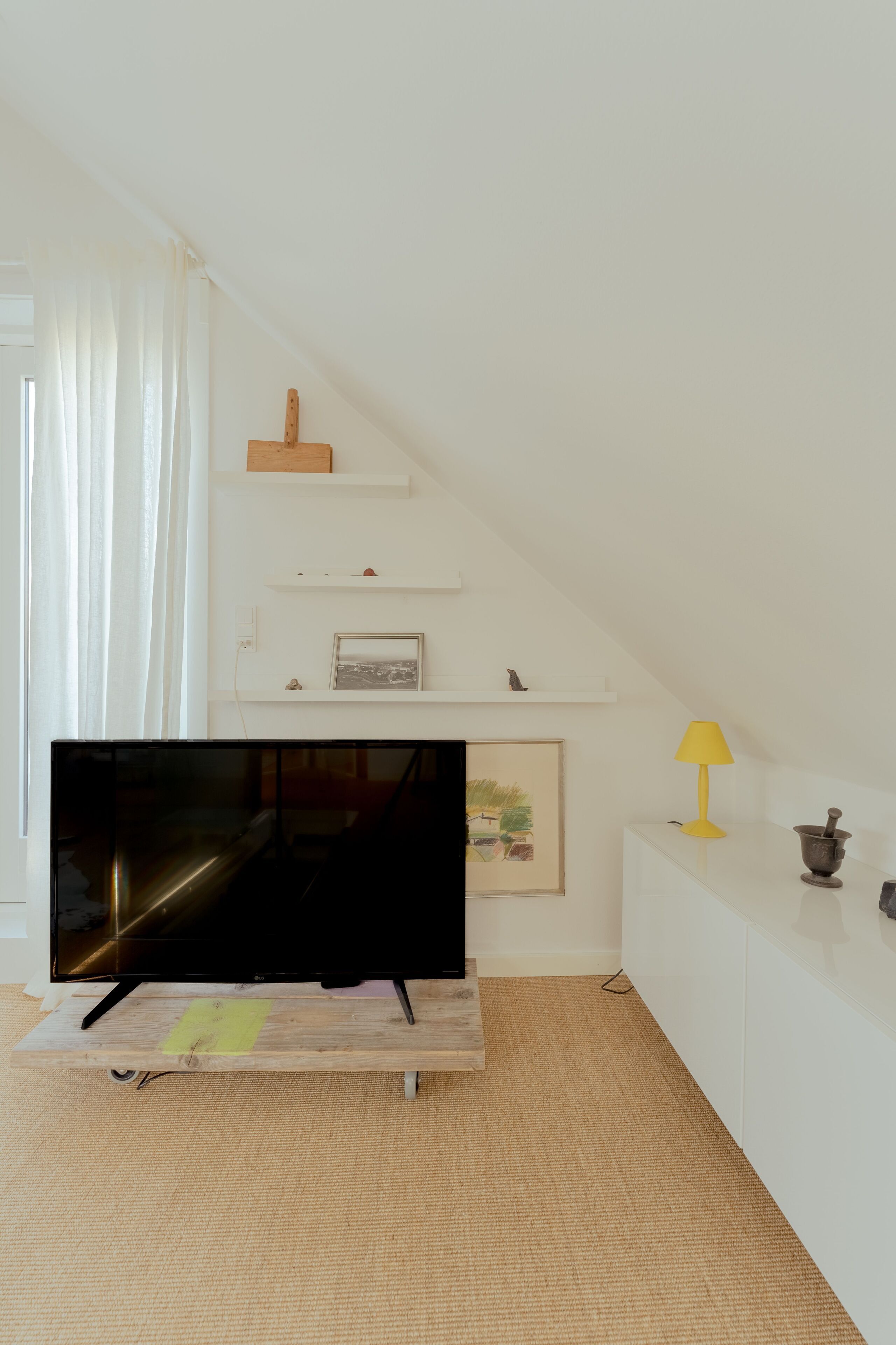 A living room with a flat-screen TV on a wooden stand, white shelves with decorative items, a yellow lamp, and a white console table.