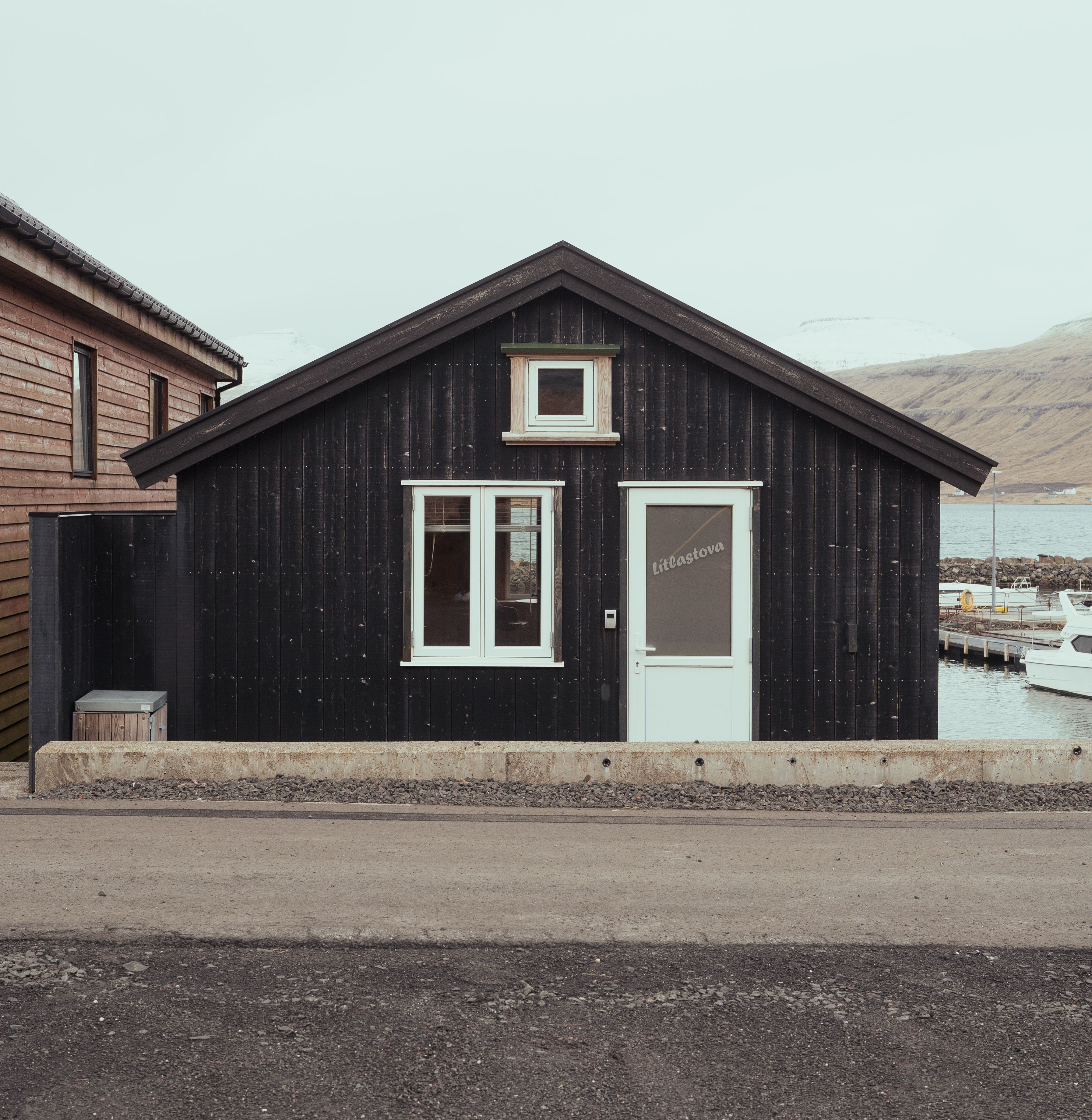 A small, black wooden building with a white door and windows, located near a body of water.
