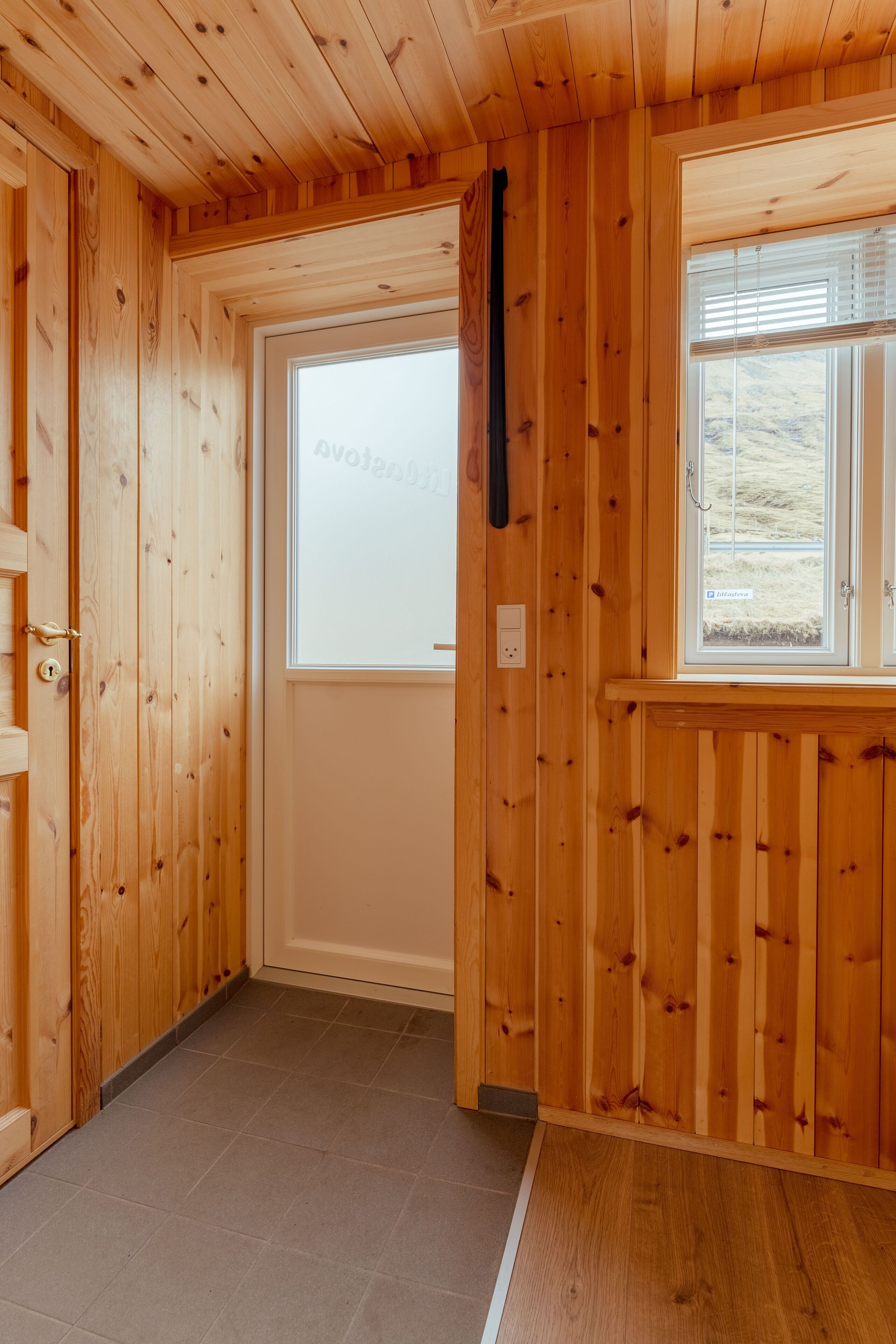 A wooden interior hallway with a door leading to a room, a window with blinds, and a light switch on the wall.