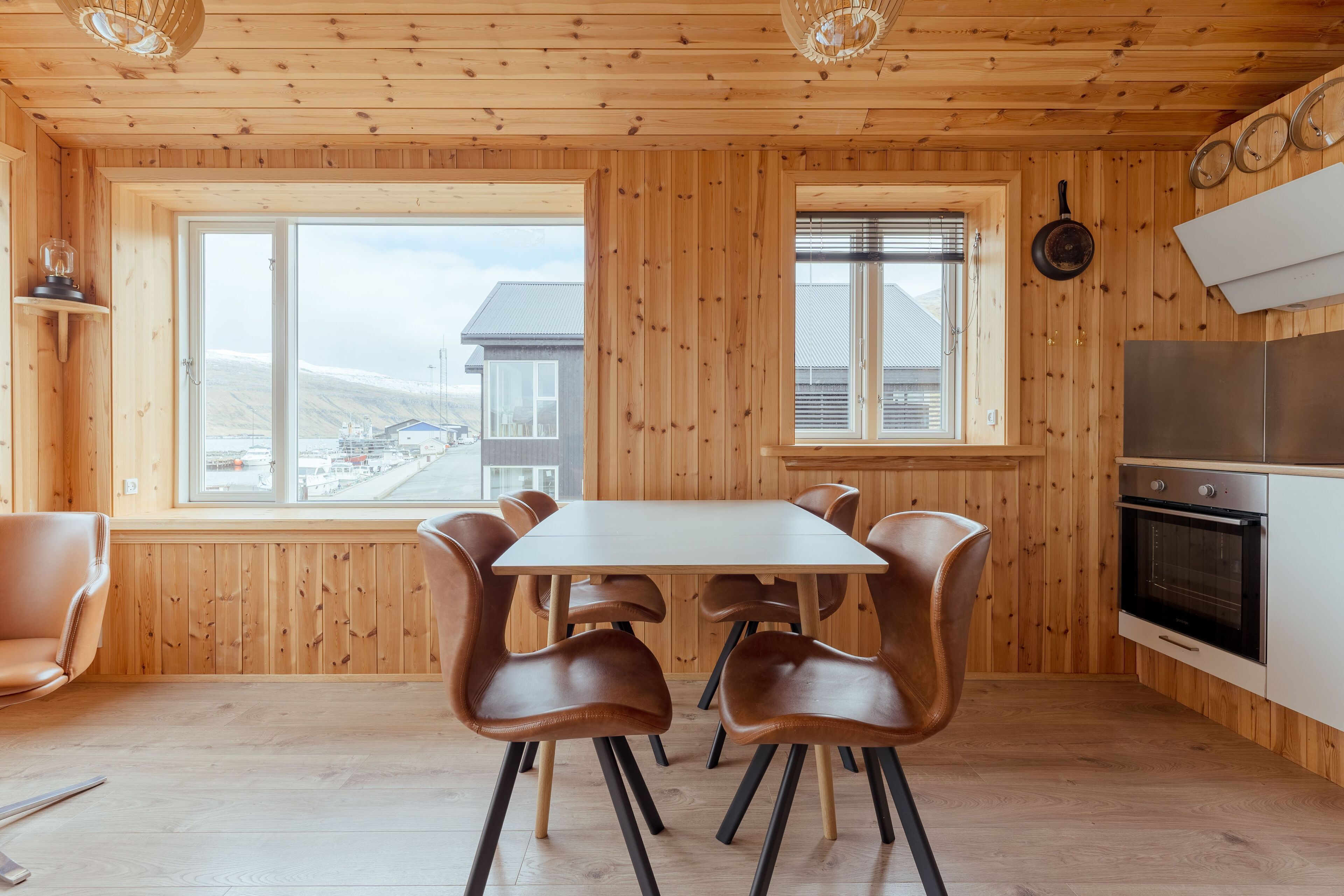 Une pièce aux murs lambrés, comprenant une table à manger et des chaises, un coin cuisine avec un fourneau, et de grandes fenêtres offrant une vue sur un bâtiment et un paysage.