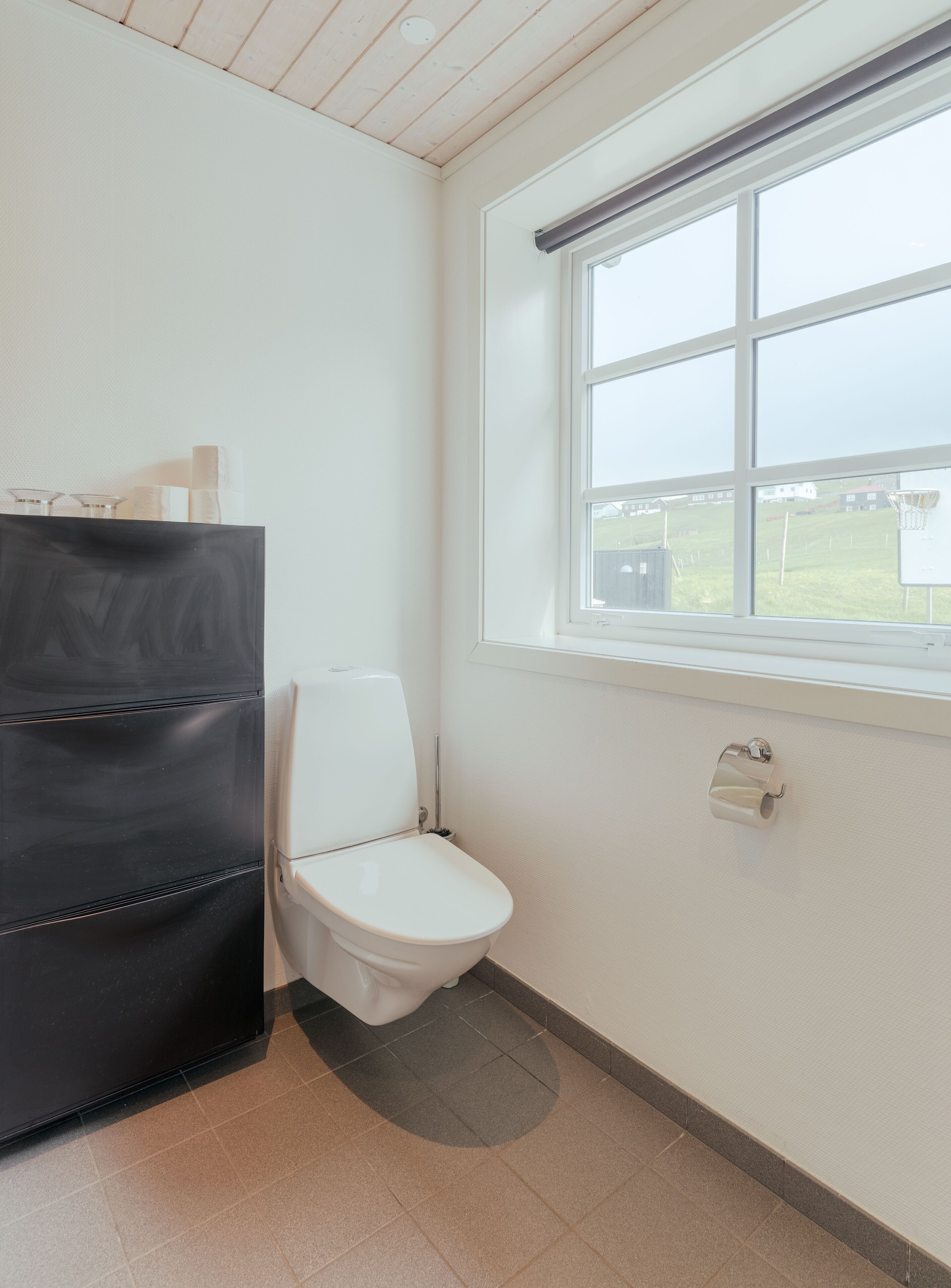 Ein kleines, sauberes Badezimmer mit Toilette, einem schwarzen Schrank und einem Fenster mit Blick auf eine Grünfläche.