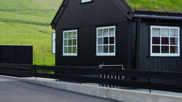 A black house with white windows, a grassy hill behind it, and a road in the foreground. - Charming Cottage In Tranquil Village - Hot Tub (Skalavik)