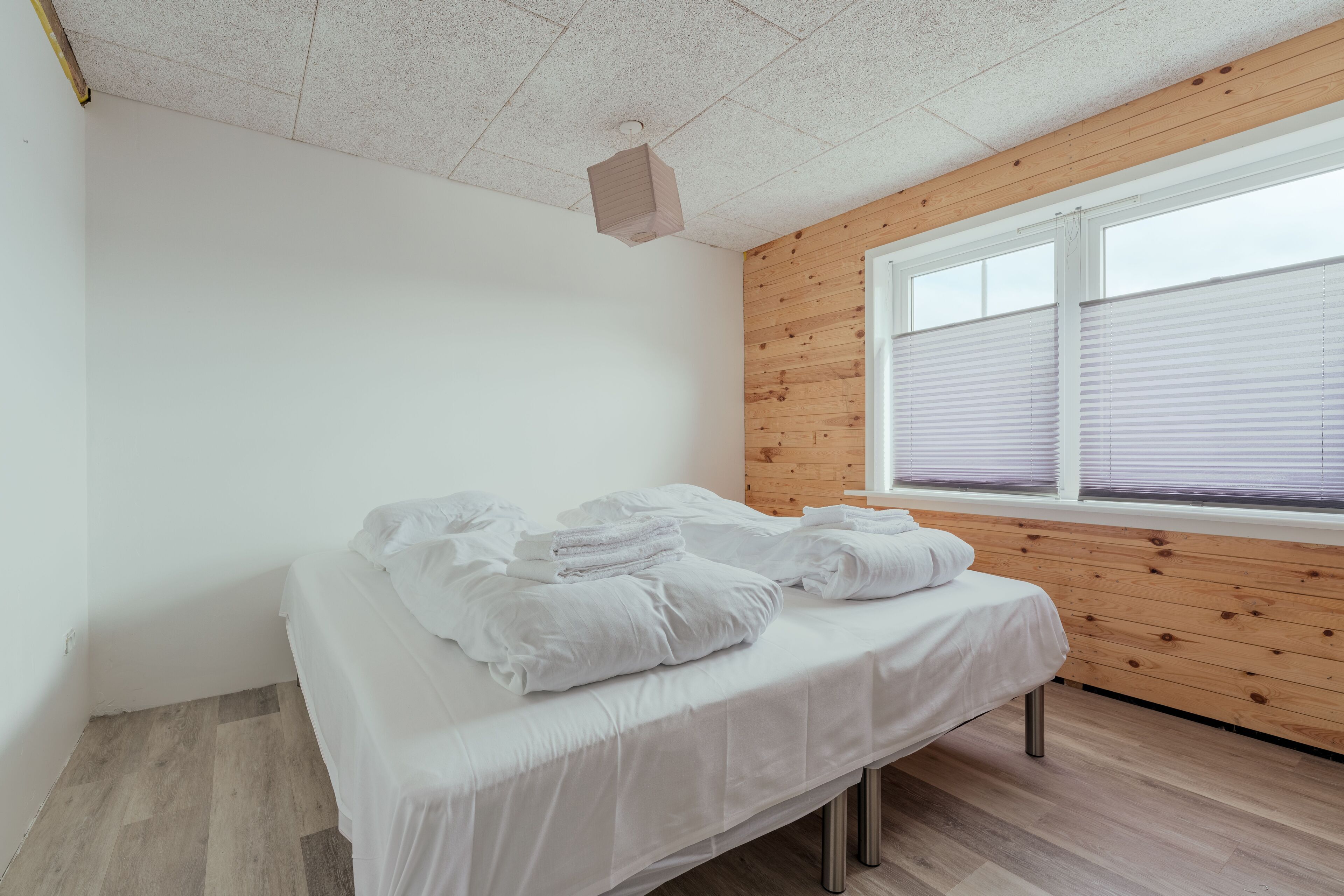 Un dormitorio con pared de madera, una ventana con persianas y una cama con ropa blanca.
