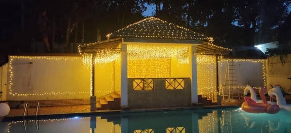A poolside gazebo with string lights, a ladder, and inflatable rings.