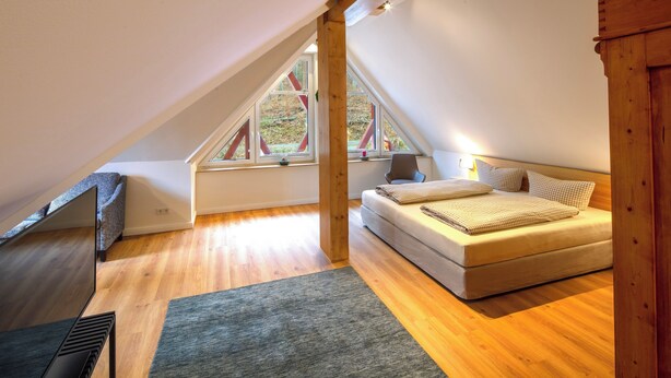 Ein Schlafzimmer mit schrägen Wänden, Holzbalken, einem Bett, einem Fenster und einer Heizung.
