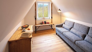Ein gemütliches Schlafzimmer im Loft-Stil mit schrägen Wänden, einem großen Fenster mit Blick auf Bäume, einem Holztisch mit Büchern und Obst sowie einem grauen Sofa.