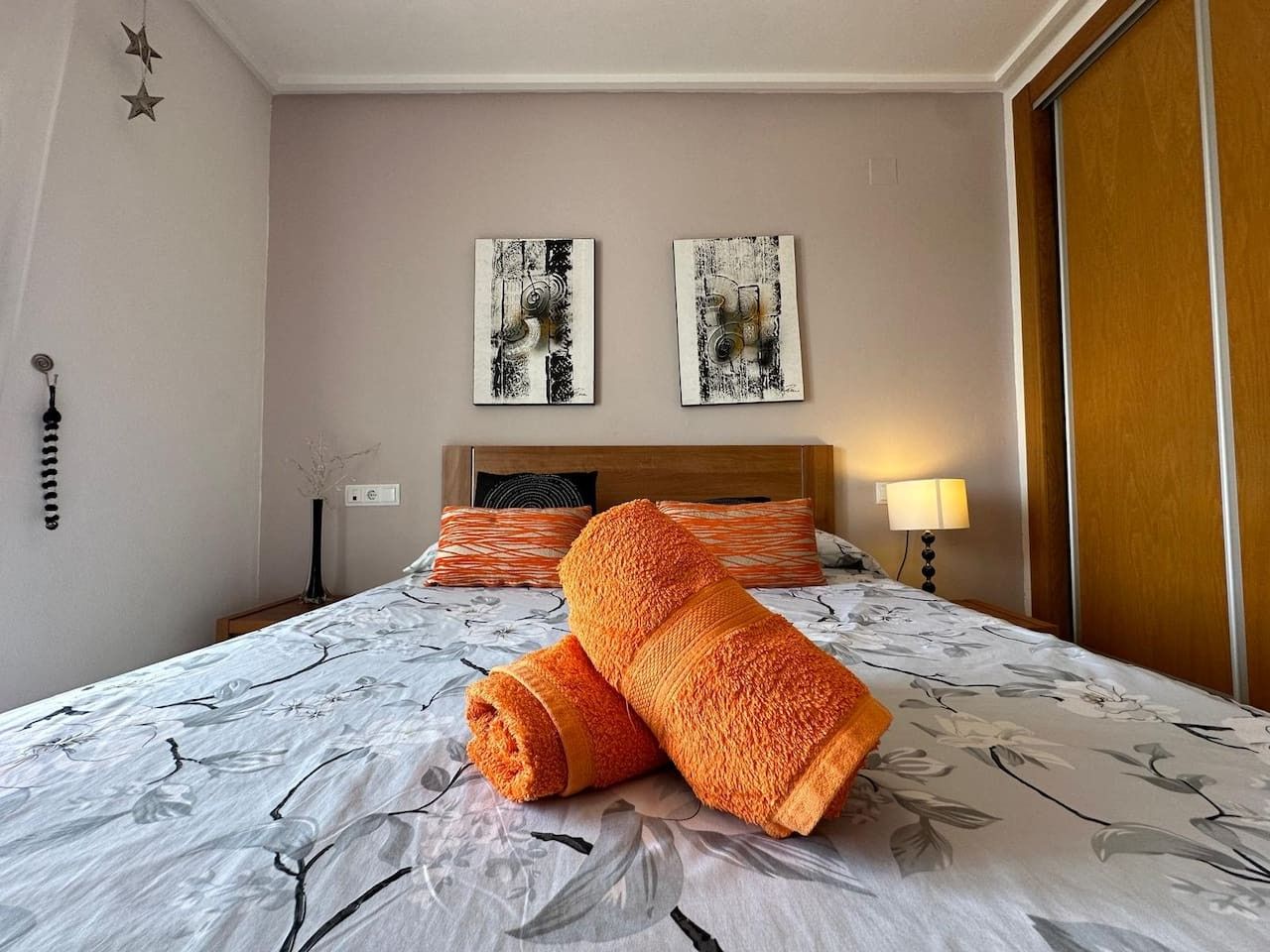 Een hotelkamer met een bed, twee kunstwerken aan de muur, een nachtkastje met een lamp, en een houten deur.