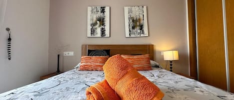 A hotel room with a bed, two wall-mounted artworks, a nightstand with a lamp, and a wooden door.