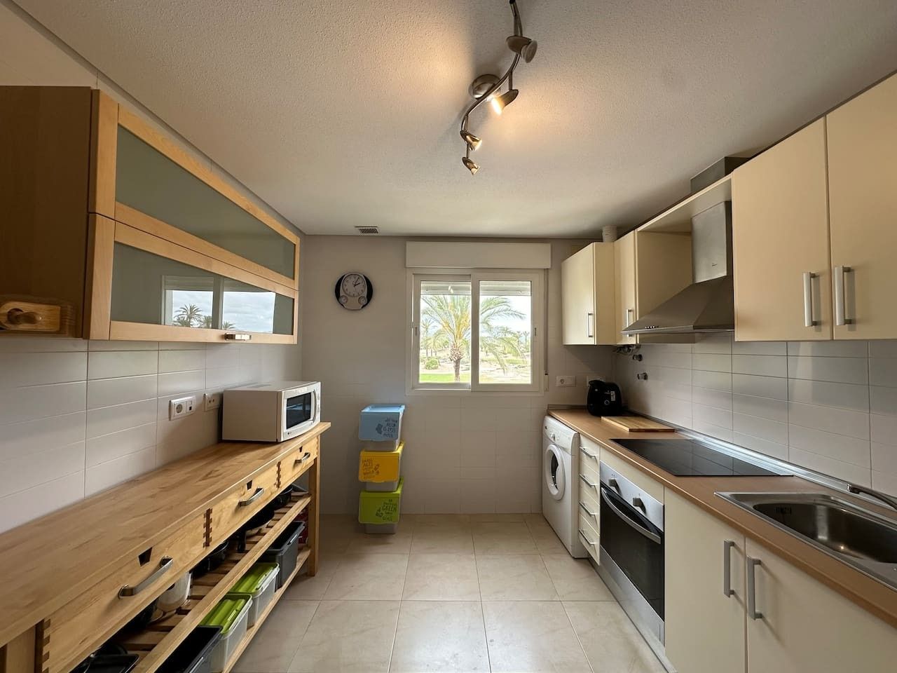 Cocina compacta equipada con microondas, lavadora y ventana con vista a palmeras.