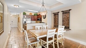Un coin repas avec une table en bois et des chaises blanches, un luminaire suspendu et une cuisine en arrière-plan.