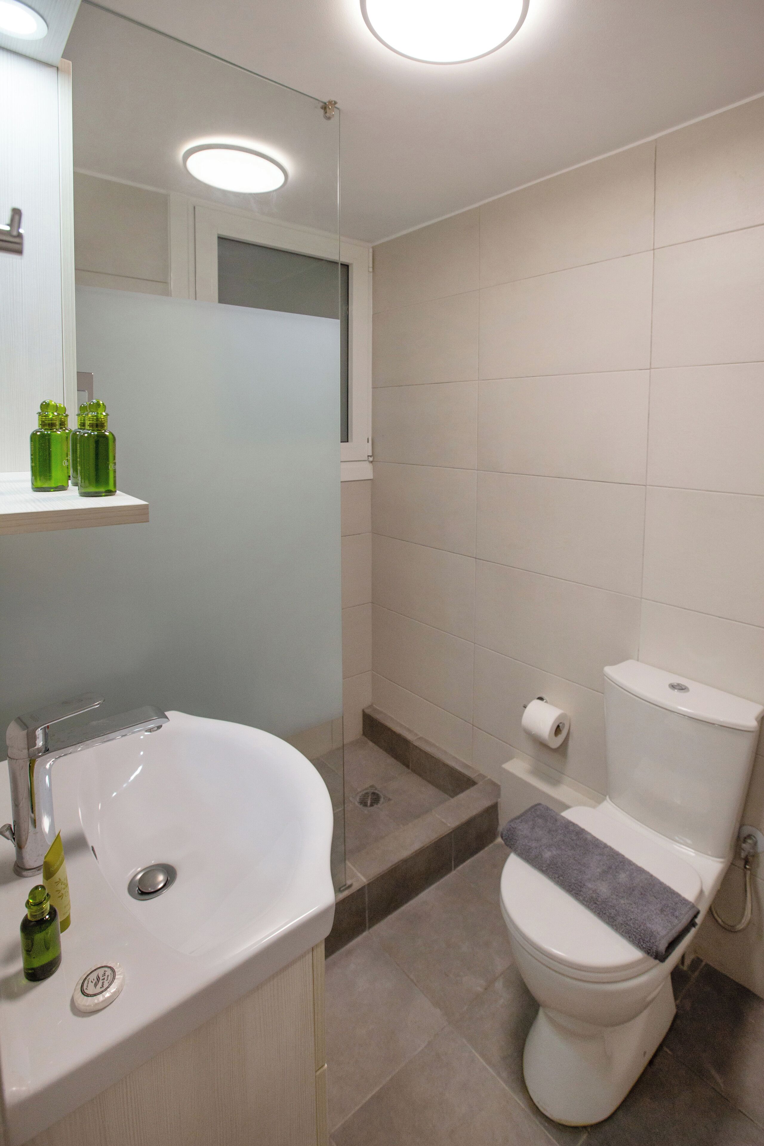 Un baño con lavamanos, espejo y retrete blancos. Hay botellas verdes sobre el lavamanos y una toalla en el retrete.