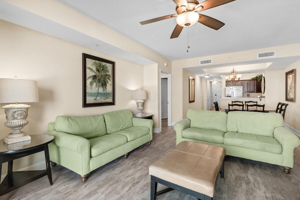 A living room with two green sofas, a beige ottoman, and a ceiling fan.