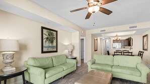 A living room with two green sofas, a beige ottoman, and a ceiling fan. - Shoreline Sunsets: 2BR Condo with Modern Comforts (North Myrtle Beach)