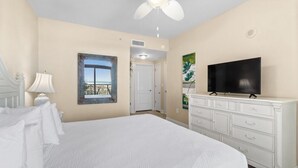 A bedroom with a large bed, a ceiling fan, a television on a dresser, and a door leading to another room. - Shoreline Sunsets: 2BR Condo with Modern Comforts (North Myrtle Beach)