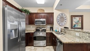 A modern kitchen with stainless steel appliances, dark wooden cabinets, a granite countertop, and a large wall clock. - Shoreline Sunsets: 2BR Condo with Modern Comforts (North Myrtle Beach)