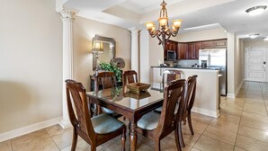 Un comedor con una mesa de vidrio rodeada de sillas de madera, una araña y una cocina al fondo.