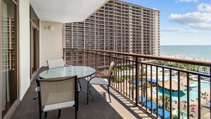 Un balcón con una mesa y sillas de vidrio con vista a una piscina y un edificio de gran altura.