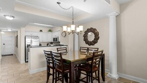 A dining area with a glass-top table surrounded by wooden chairs, a chandelier, and a decorative wreath on the wall.