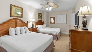 A hotel room with two beds, a wooden headboard, a ceiling fan, a desk with a lamp, and artwork on the walls.