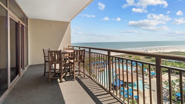 A balcony with a table and chairs, overlooking a beach and a pool area.