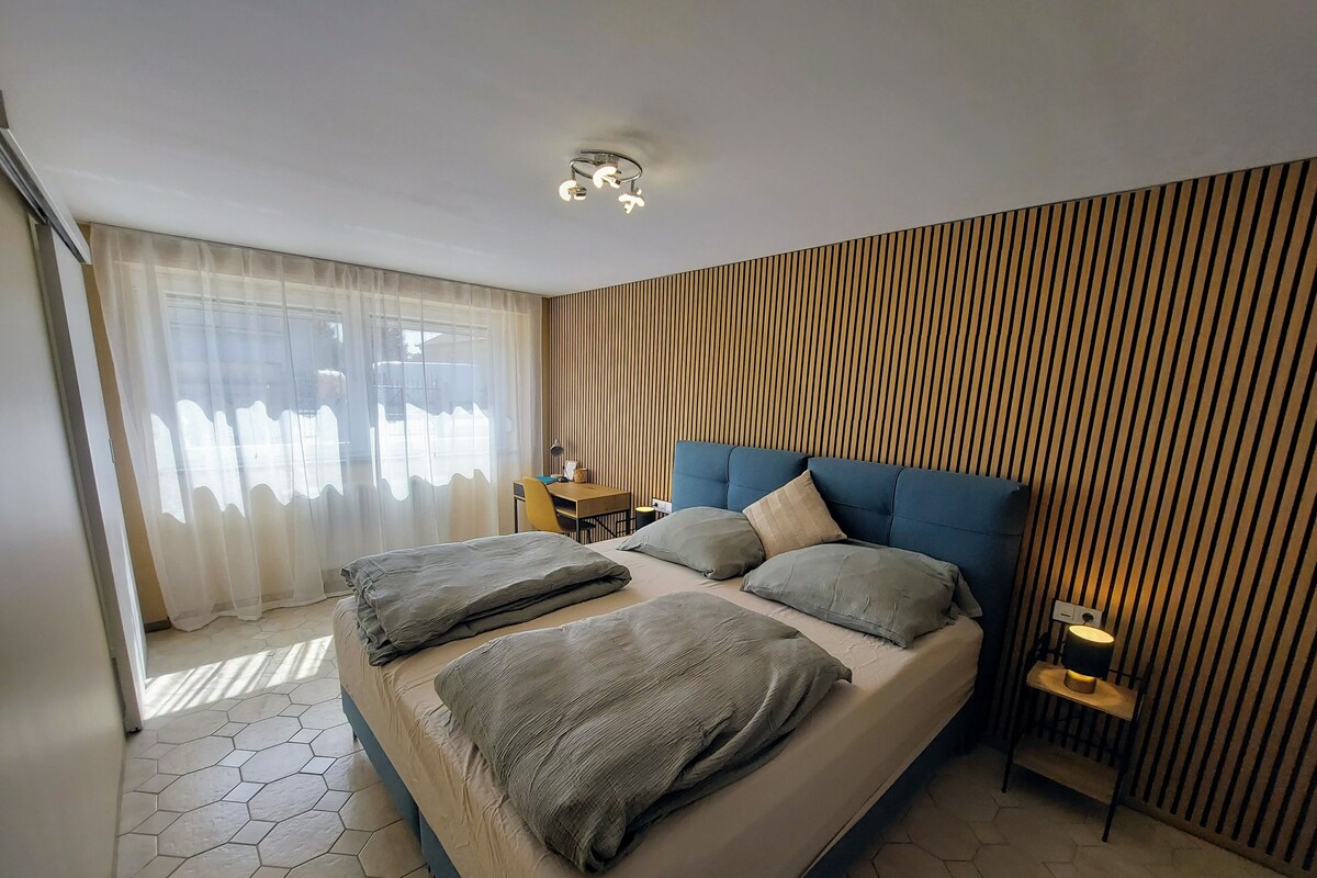 Une chambre moderne avec un grand lit, un mur lambrissé en bois, un bureau avec une chaise et une grande fenêtre ornée de rideaux légers.