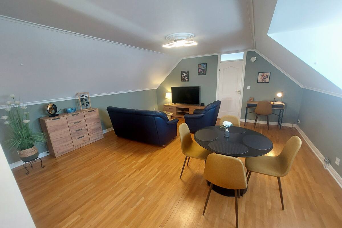 Ein Lofts-Wohnraum mit hohen Decken, Holzböden, einem dunkelblauen Sofa, einem runden Esstisch mit Stühlen, einem Fernseher und einem kleinen Arbeitsbereich mit Schreibtisch und Stuhl.