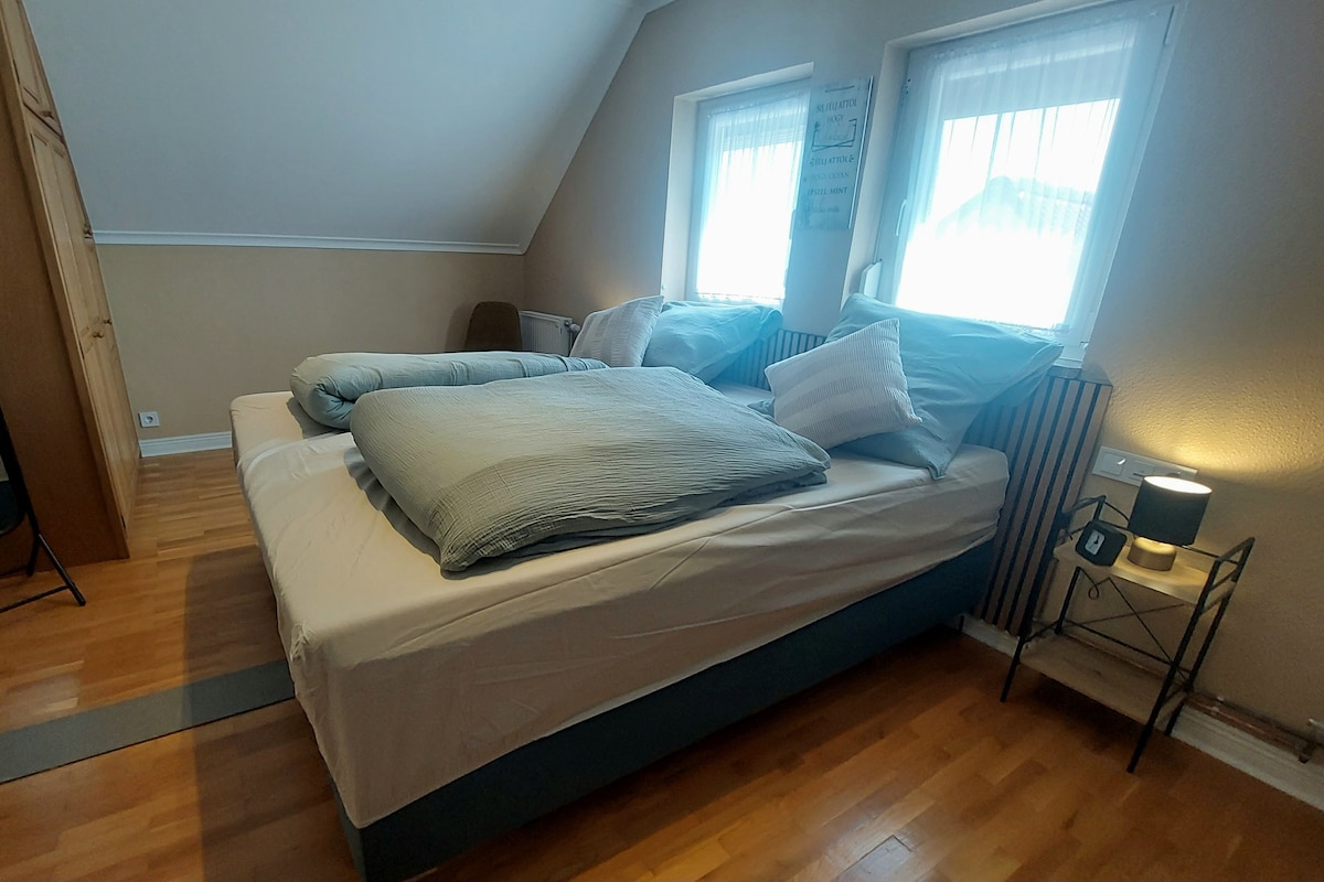 Un dormitorio con cama, mesita de noche con lámpara, ventana con cortinas y un espejo.