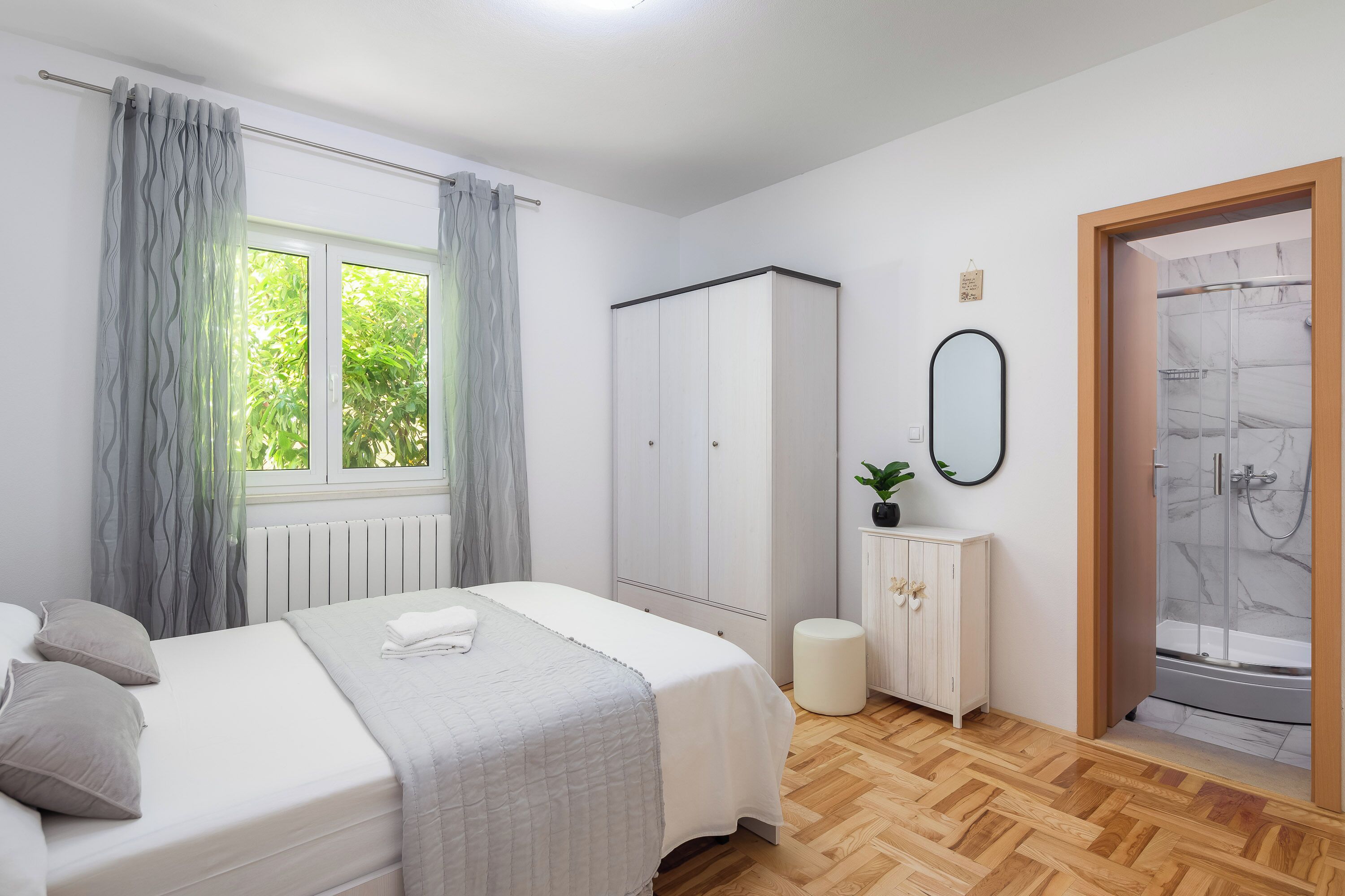 A bedroom with a bed, a wardrobe, a radiator, a small stool, a cabinet, a mirror, and a shower area.