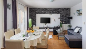 Ein modernes Wohnzimmer mit einem an einer Ziegelwand montierten Flachbildfernseher, einem dunkelblauen Sofa und einem Essbereich mit Tisch für vier Personen.