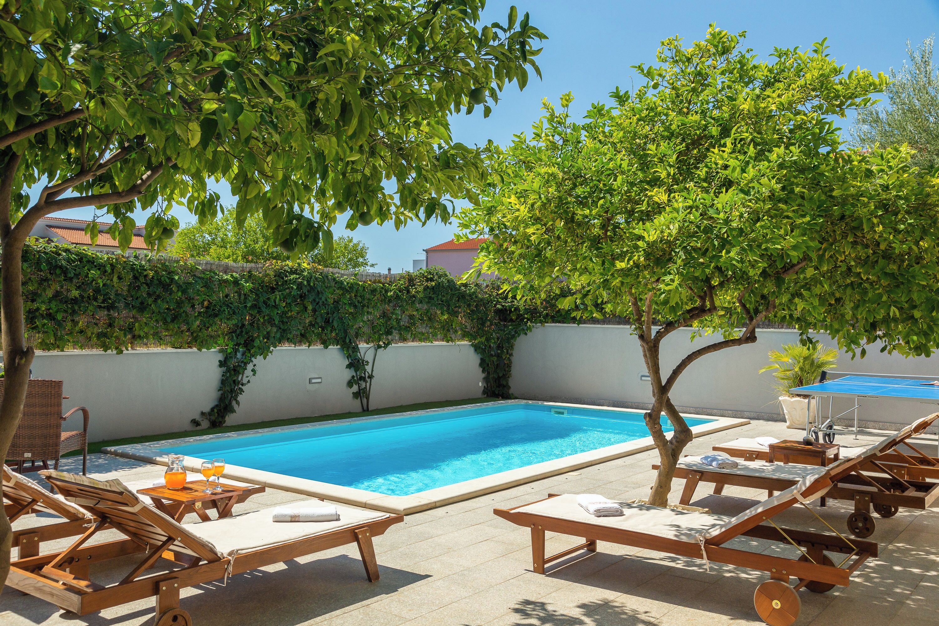 Piscina con sillones de descanso y una mesa, rodeada de árboles y setos.