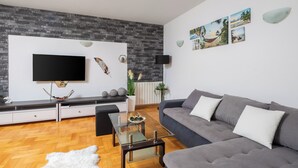 Ein modernes Wohnzimmer mit Flachbildfernseher an einer Wand mit Steinoptik, einem grauen Sofa, einem Glastisch und Holzboden.