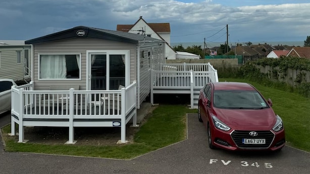 Une maison mobile dotée d’une terrasse clôturée et d’une voiture rouge garée à proximité.