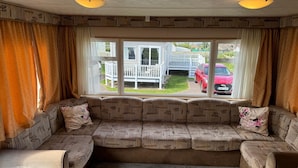 Un salon chaleureux avec une grande fenêtre, un canapé d’angle et une vue sur une maison blanche et une voiture rouge garée à l’extérieur.