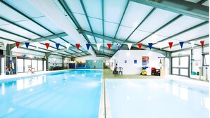 Une piscine intérieure avec un plafond haut et des poutres métalliques. Des drapeaux colorés sont suspendus au plafond, et on peut apercevoir quelques chaises près de la piscine.
